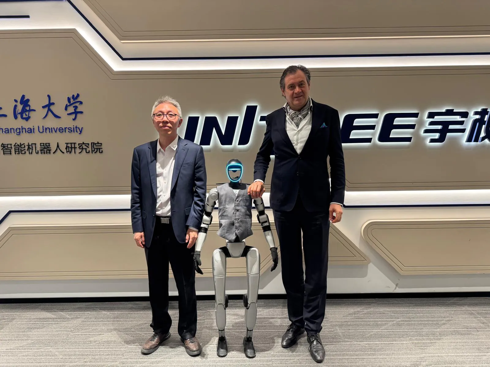 Asinov's Benoît Dubuis and Professor Chen of Shanghai University standing with a Unitree humanoid robot, beneath the Shanghai University and Unitree signage