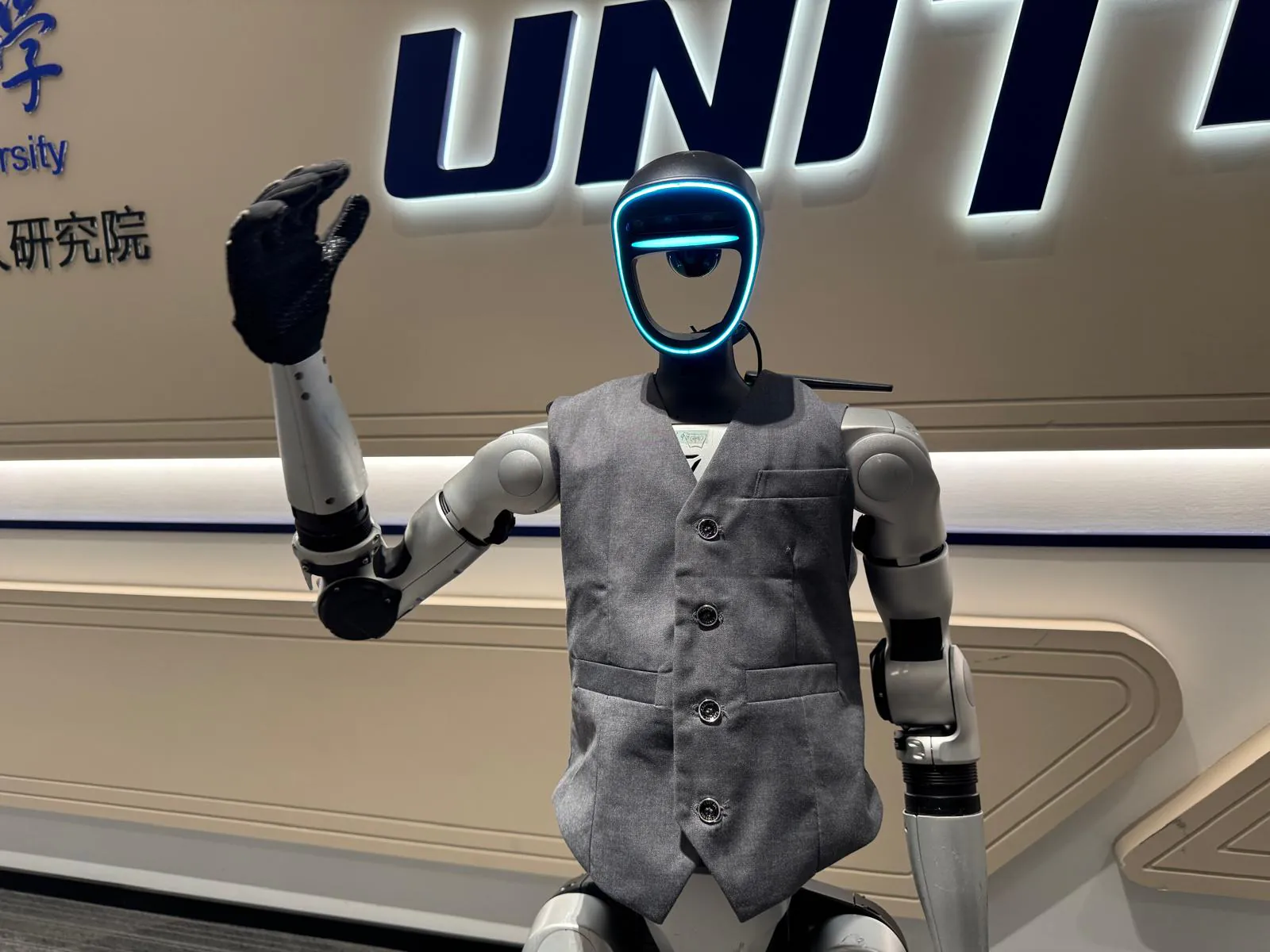 A Unitree humanoid robot in a gray vest raises a hand in greeting, in front of the Unitree logo